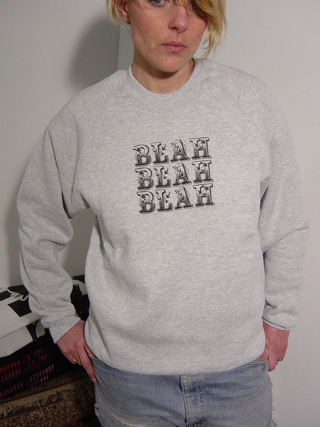 A person wearing a light heather gray crewneck sweatshirt with 'BLEAH BLEAH BLEAH' printed in a black-and-white block pattern on the chest, paired with light blue jeans.