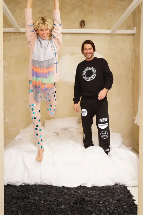 Two adults in colorful, playful loungewear standing and jumping on a bed in a cozy beige room.