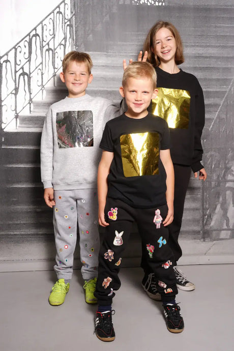 Three kids wearing metallic square print shirts and playful patchwork sweatpants, posing indoors.