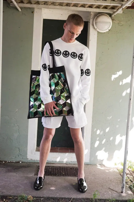 Person wearing white smiley face sweatshirt and shorts, holding large geometric tote bag