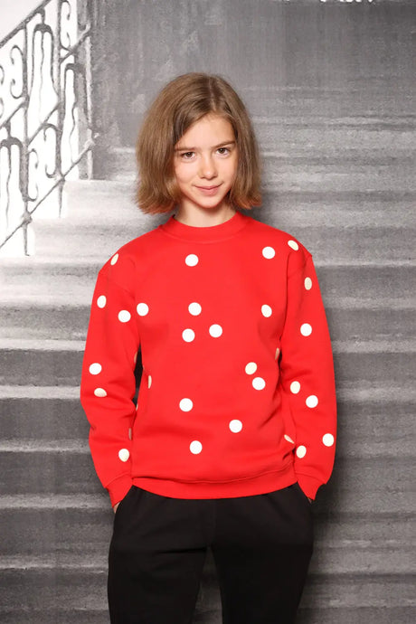 Child wearing red sweater with white polka dots, standing against gray staircase background