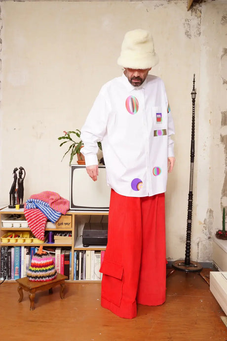 Man wearing white gradient print shirt, red wide-leg pants, and cream hat indoors