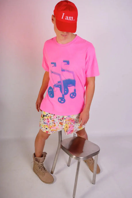 Person in red 'I am.' cap, pink graphic t-shirt, colorful shorts, tan boots, studio setting