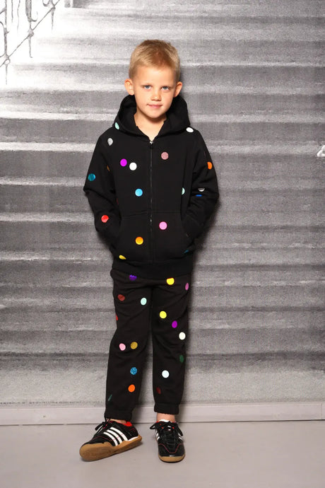 Child in black jogger set with colorful polka dots, standing against a gray background