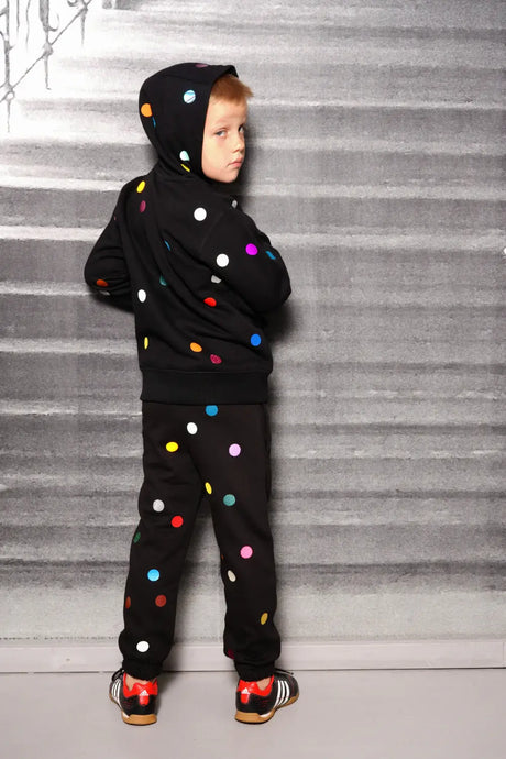 Child in black hoodie and pants with colorful polka dots, standing against a gray wall