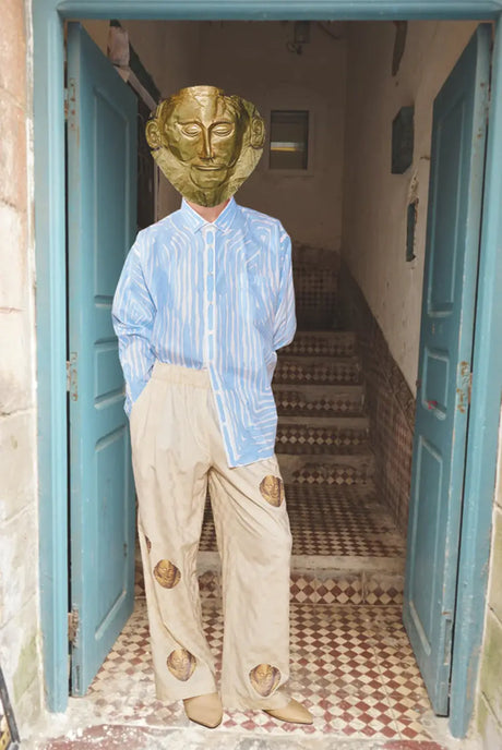 Mannequin in blue striped shirt, beige pants with face prints, golden mask, standing in doorway