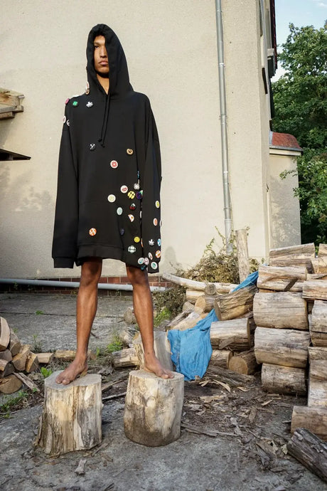 Model in oversized black hoodie with colorful patches, standing barefoot on logs outdoors