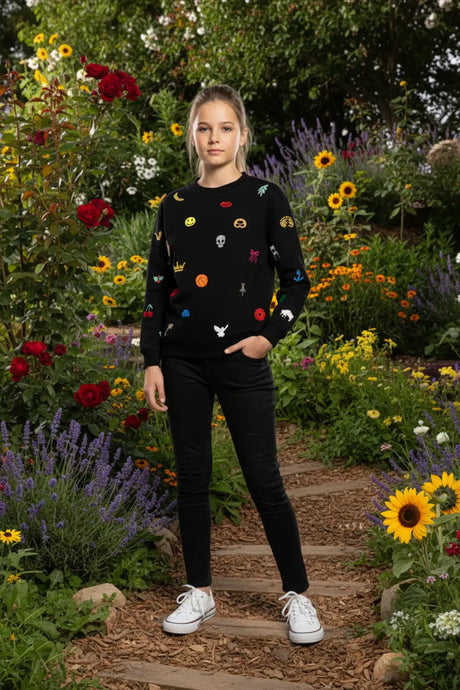 Girl in a black sweater with colorful embroidered icons, standing in a vibrant flower garden.
